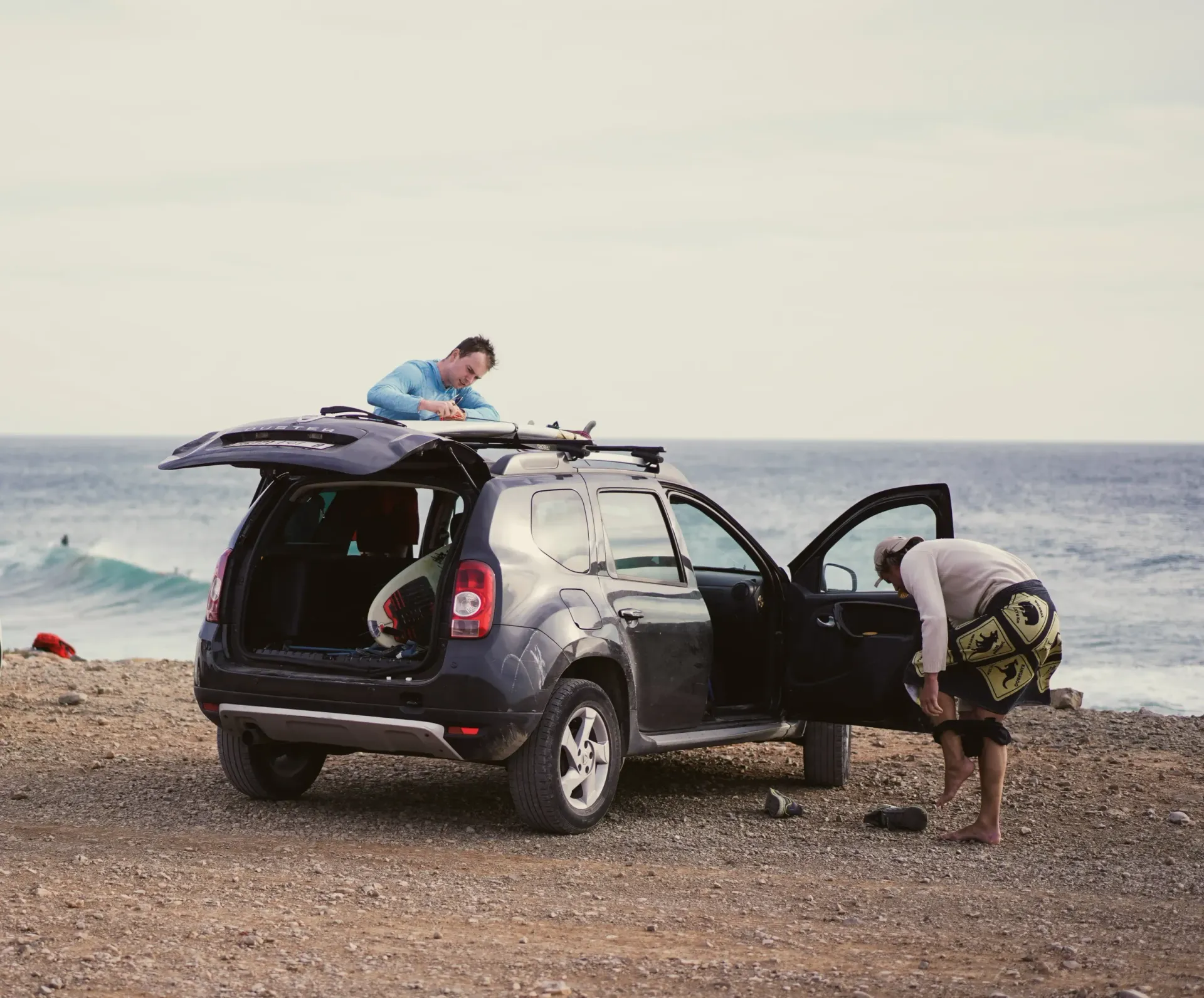 Two surfers prepare gear by a car near the ocean in Imsouane.