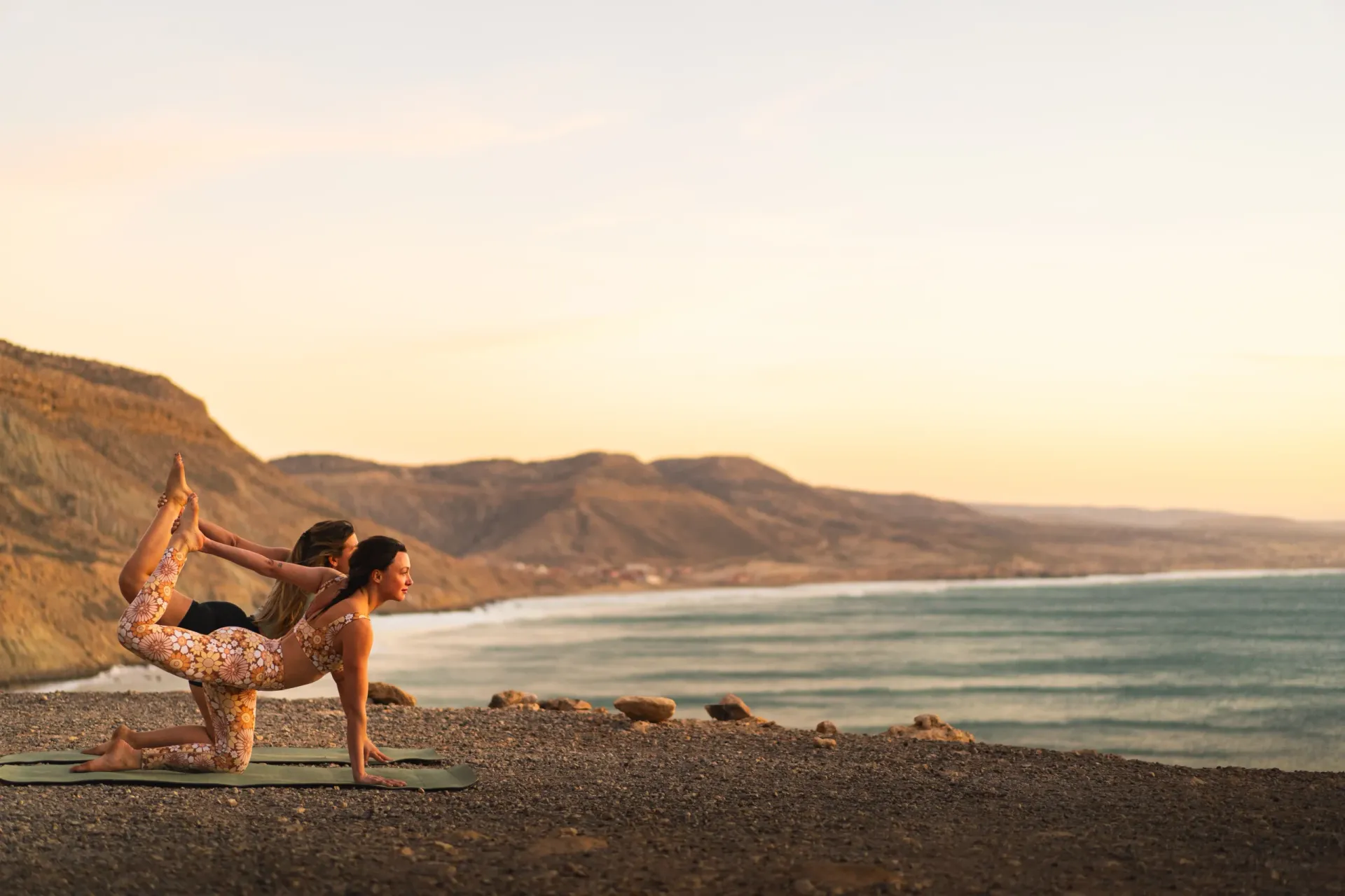 Two people practicing yoga on a cliff overlooking the ocean at sunset.